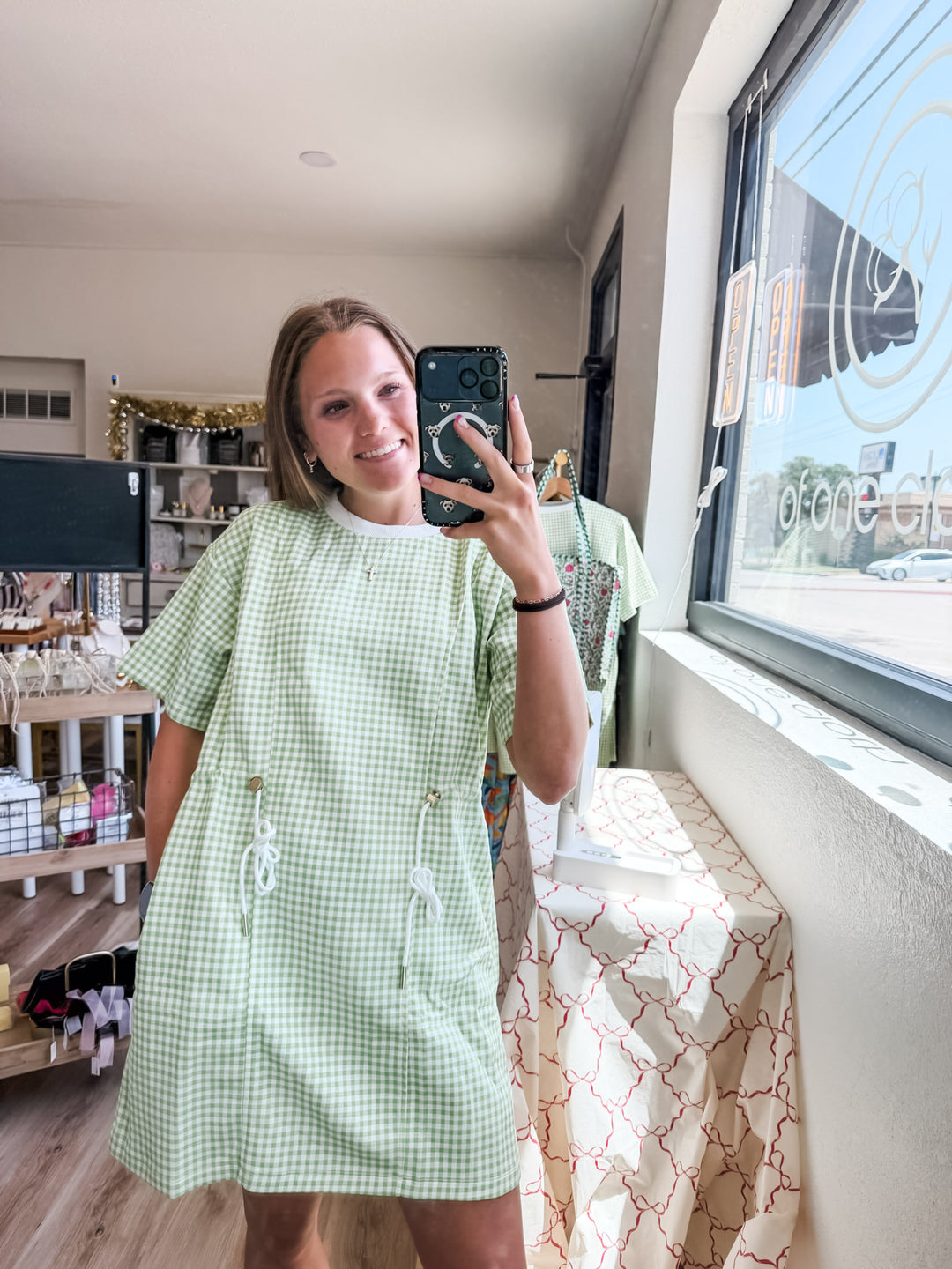 A woman takes a mirror selfie in a boutique wearing a light green and white gingham short-sleeve dress with white rope details. The shop interior is visible in the background, including shelves, a window, and a patterned table runner.