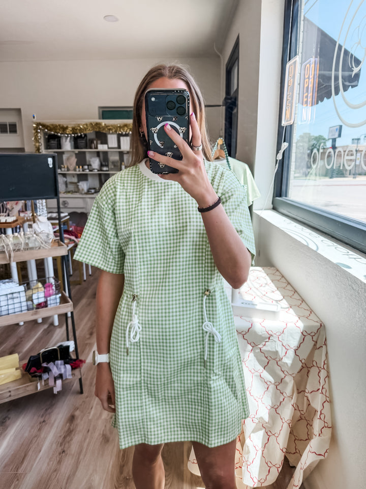 A woman takes a mirror selfie wearing a light green and white gingham short-sleeve dress in a retail store setting.