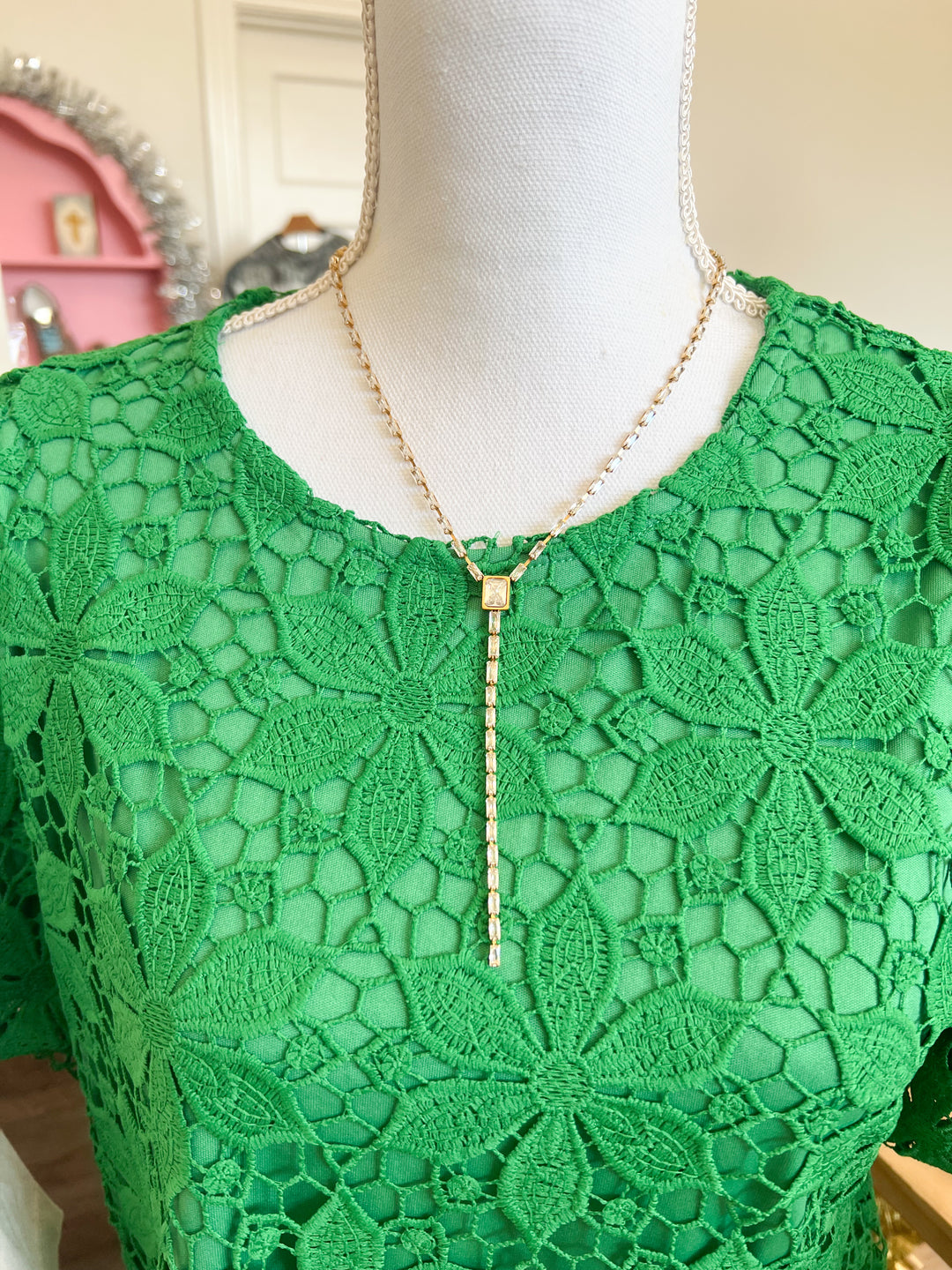 A gold lariat-style necklace with a beaded chain and a rectangular pendant, displayed on a white mannequin torso wearing a green lace top.