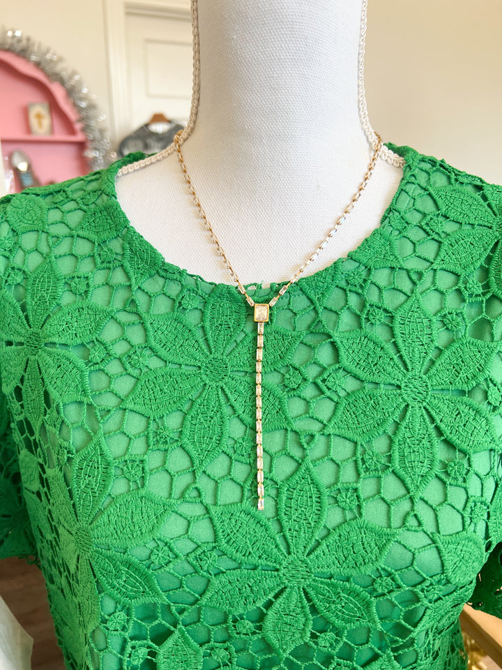 A gold lariat-style necklace with a beaded chain and a rectangular pendant, displayed on a white mannequin torso wearing a green lace top.
