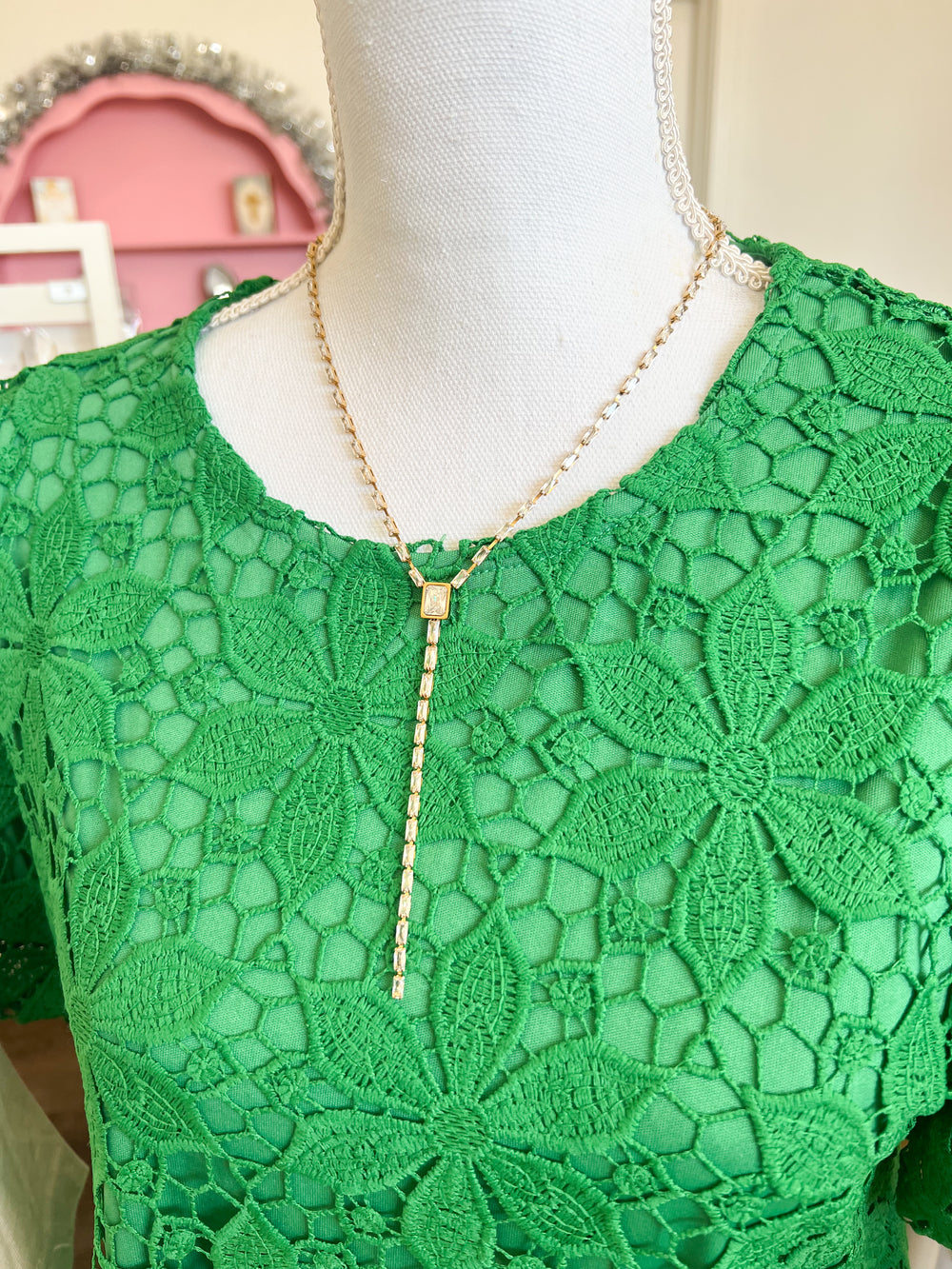 A gold-toned lariat-style necklace with a small rectangular pendant and a long drop chain, displayed on a white mannequin torso wearing a green lace top.