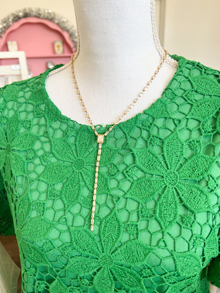 A gold-toned lariat-style necklace with a small rectangular pendant and a long drop chain, displayed on a white mannequin torso wearing a green lace top.