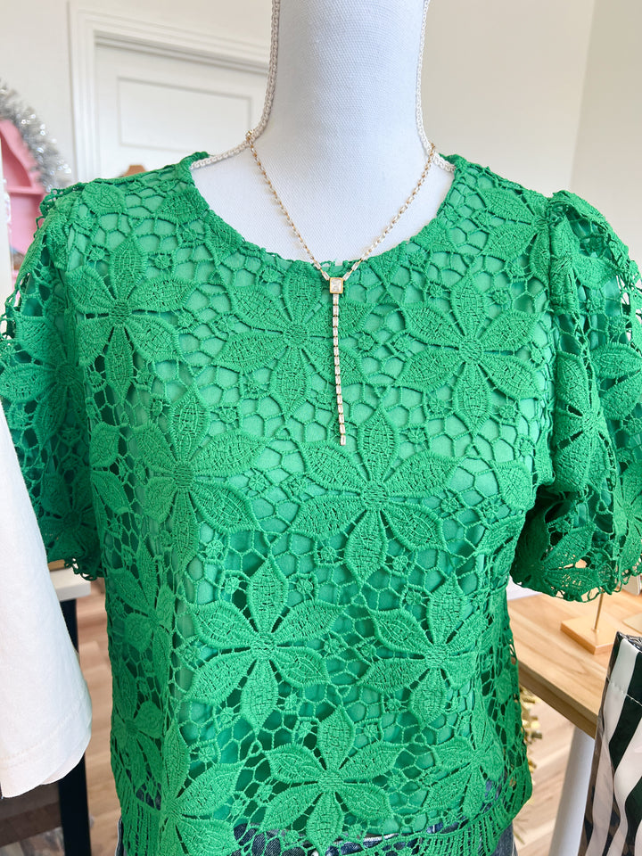 A bright green floral lace short-sleeve top displayed on a white mannequin torso, styled with a gold beaded necklace.