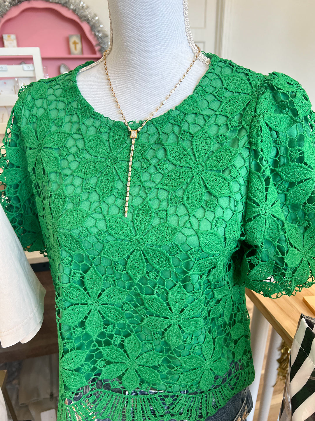 A bright green floral crochet lace top displayed on a mannequin torso, featuring short puff sleeves and a round neckline, with a gold-toned necklace draped over it.