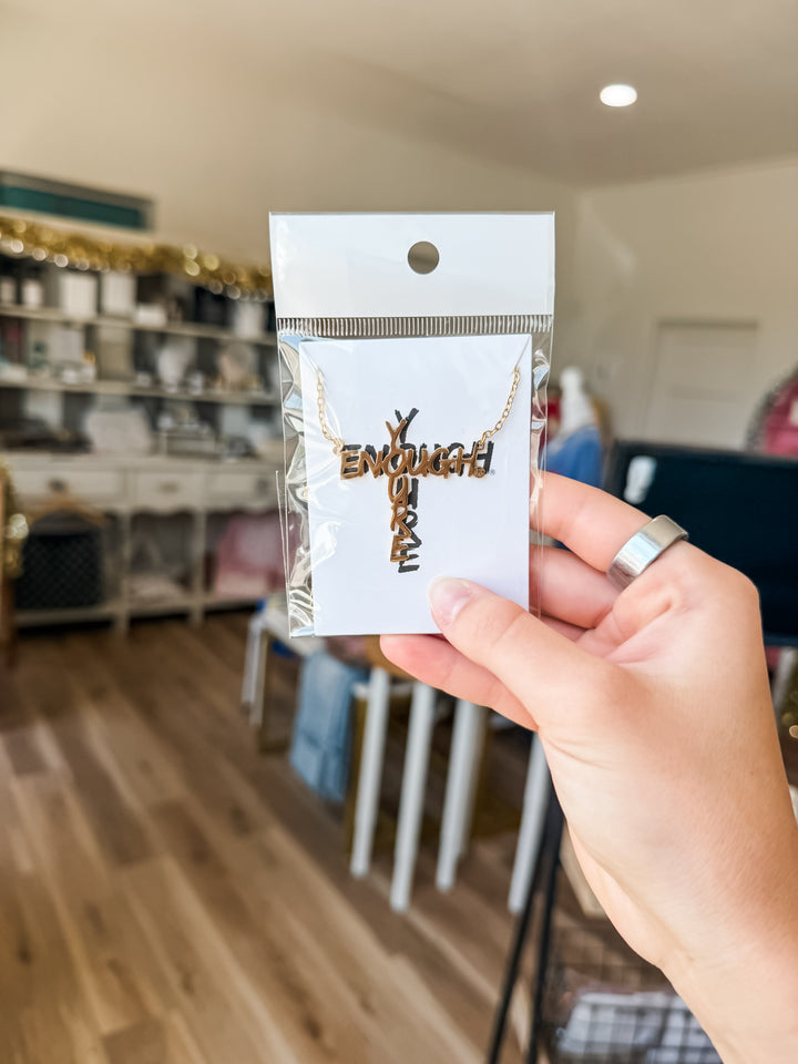 A hand wearing a silver ring holds a packaged gold necklace with a cross pendant that spells &