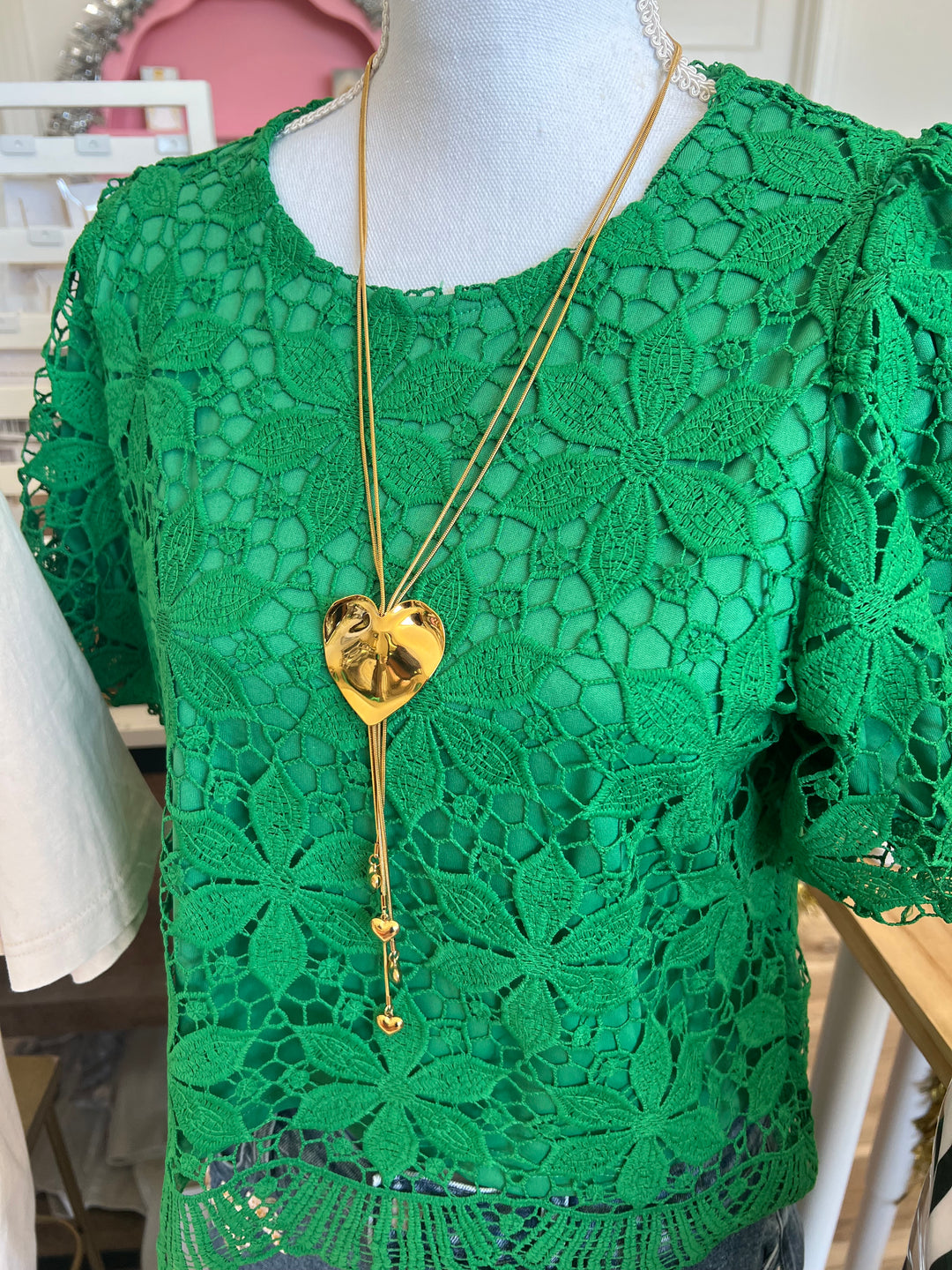A bright green floral lace short-sleeve top displayed on a mannequin torso, styled with a long gold heart pendant necklace.