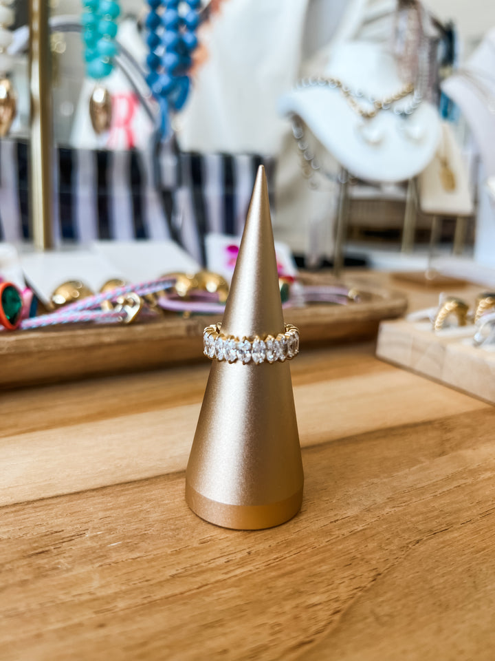 A gold ring stand displaying a gold band ring with clear gemstones, set on a wooden tabletop with a blurred jewelry store background.