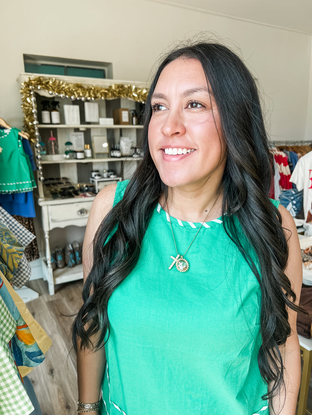 A smiling woman with long dark hair wearing a bright green sleeveless top with white trim, standing in a boutique interior with clothing racks and shelves in the background.