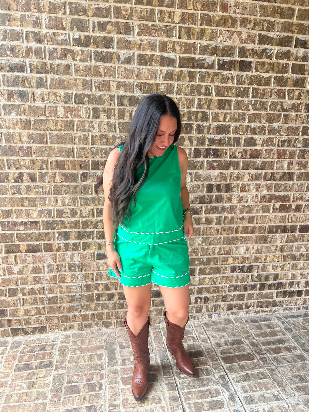 A woman wearing a bright green sleeveless top and matching green shorts with white dashed trim, paired with brown cowboy boots, standing in front of a brown brick wall.