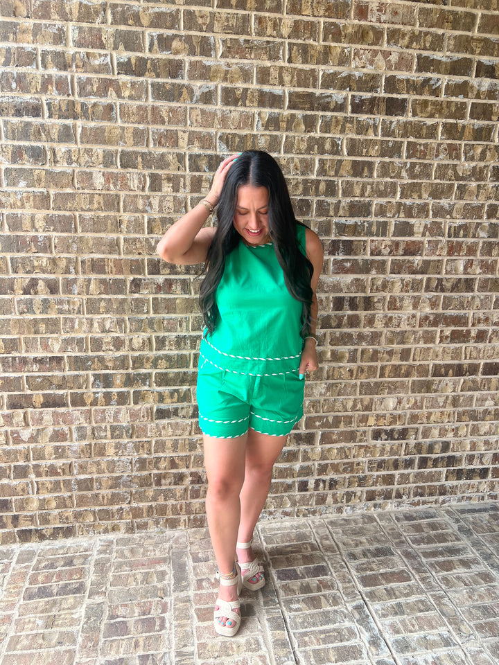 A woman wearing a bright green sleeveless top and matching green shorts with white dashed trim, standing against a brown brick wall.