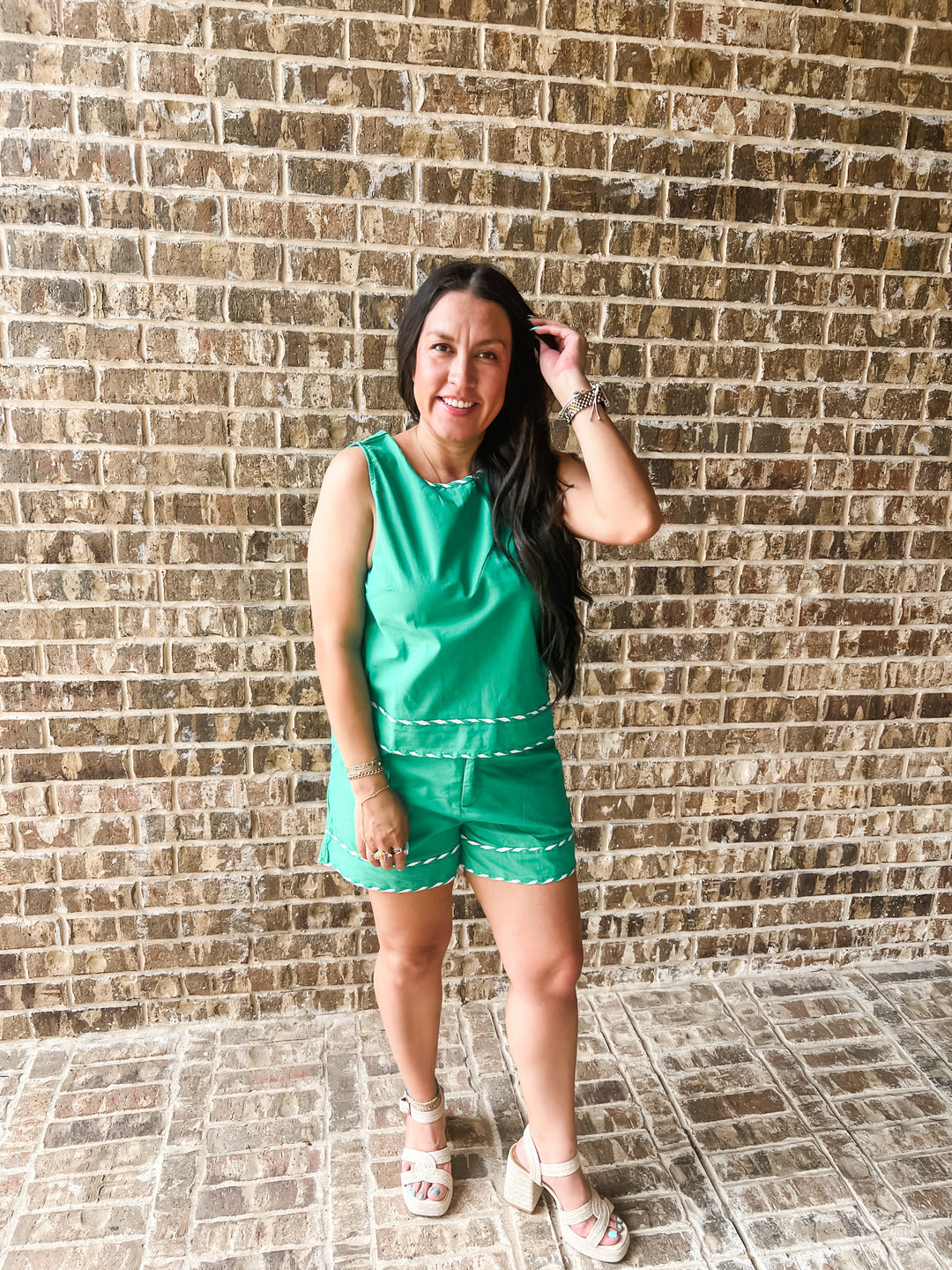 A woman modeling a bright green sleeveless romper with white trim, standing in front of a brown brick wall.