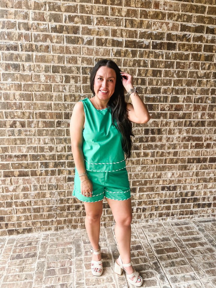A woman modeling a bright green sleeveless romper with white trim, standing in front of a brown brick wall.