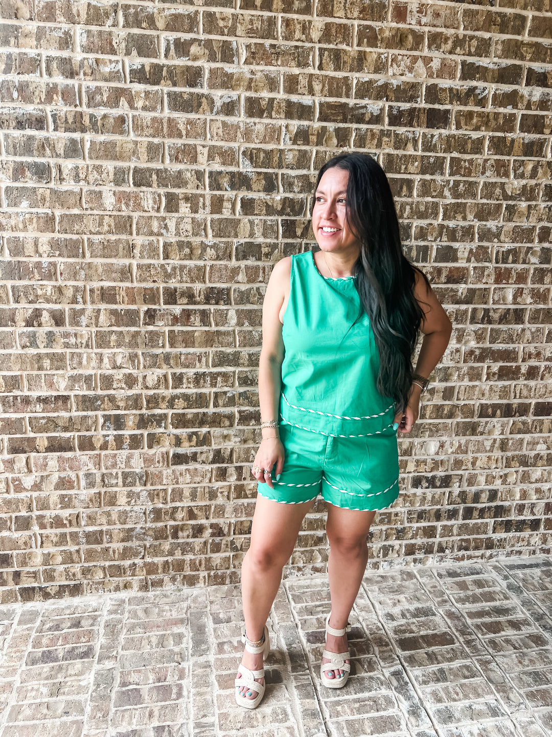 A woman modeling a bright green sleeveless romper with white contrast stitching, standing against a brown brick wall.