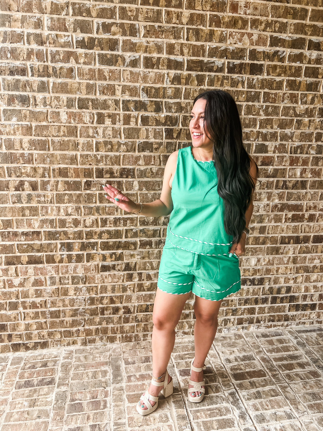 A woman wearing a bright green sleeveless romper with white contrast stitching stands in front of a brown brick wall, gesturing with her hand.