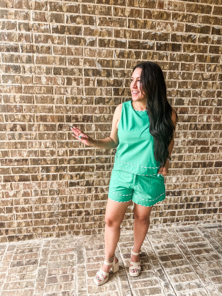A woman wearing a bright green sleeveless romper with white contrast stitching stands in front of a brown brick wall, gesturing with her hand.