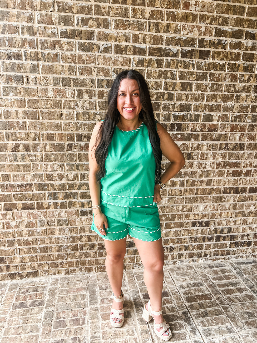 A woman modeling a bright green sleeveless romper with white contrast stitching, standing in front of a brown brick wall.