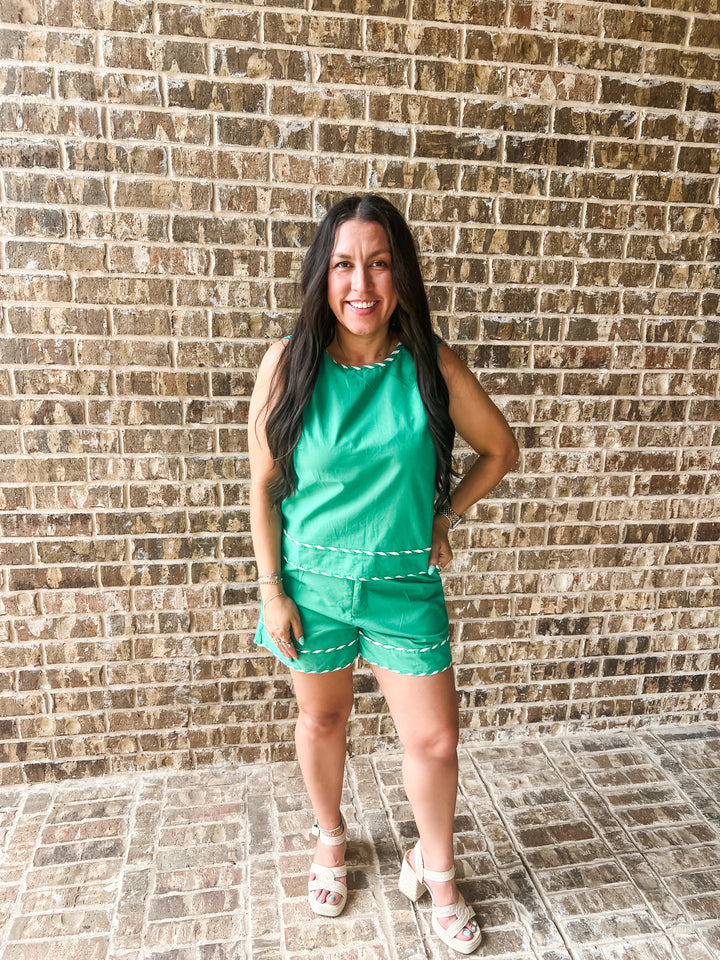 A woman modeling a bright green sleeveless romper with white contrast stitching, standing in front of a brown brick wall.