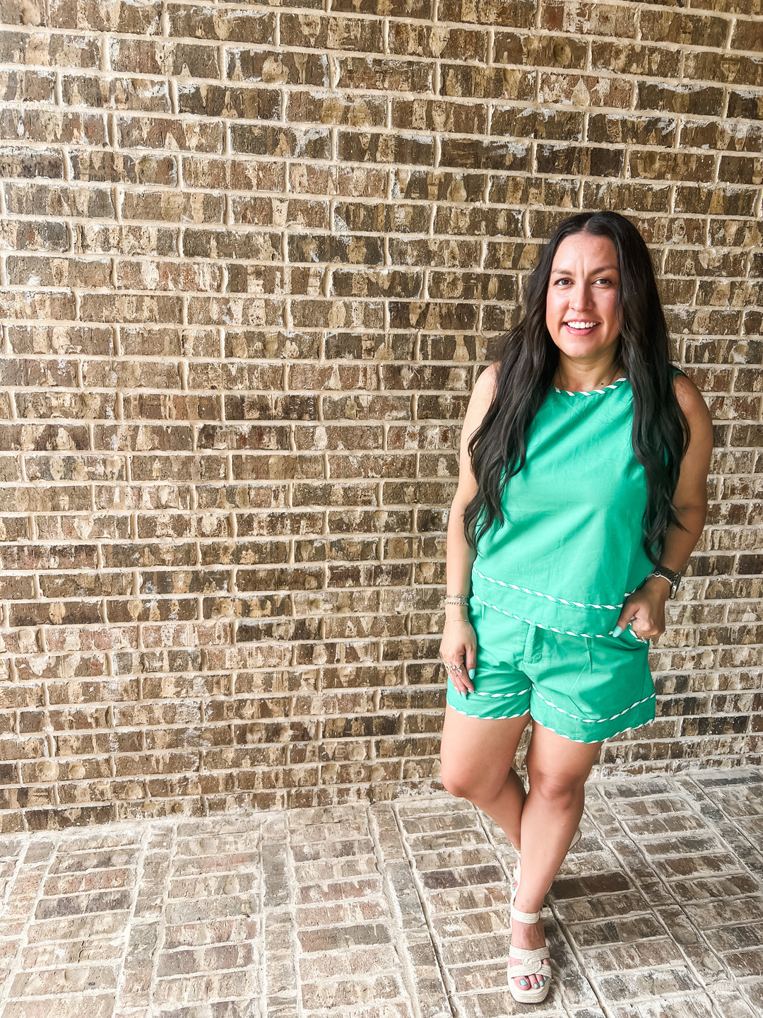 A woman posing in a bright green sleeveless top and matching shorts with white contrast stitching, standing against a brown brick wall.