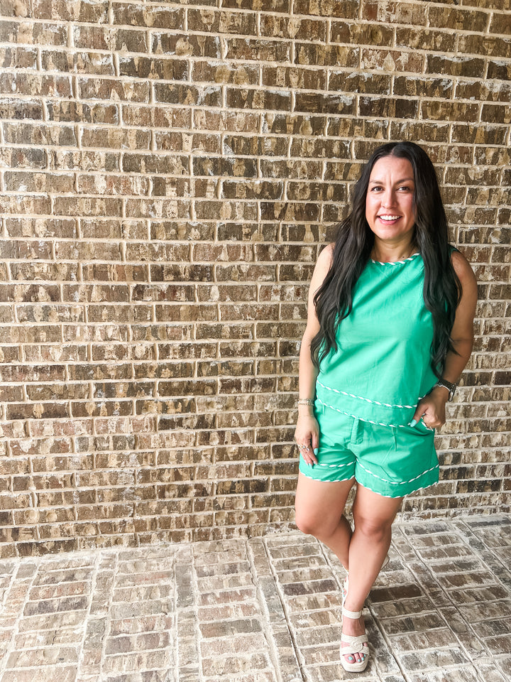 A woman posing in a bright green sleeveless top and matching shorts with white contrast stitching, standing against a brown brick wall.