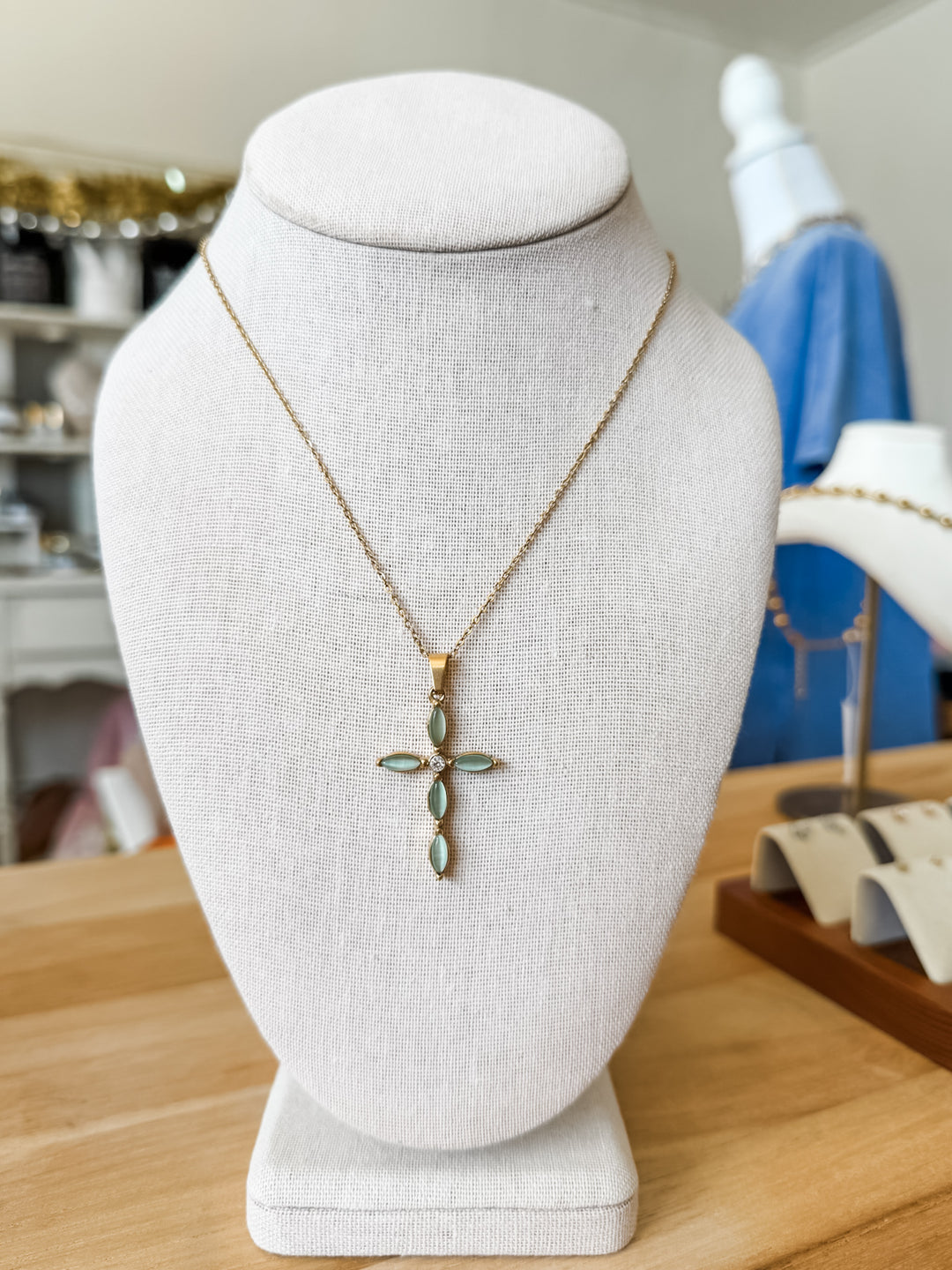 A gold cross pendant necklace with green marquise-cut stones, displayed on a white fabric jewelry bust.