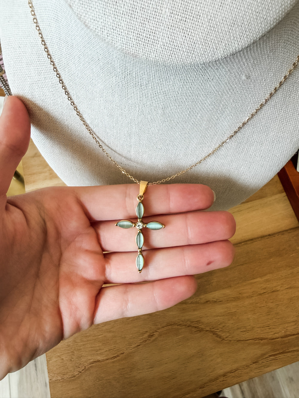 A gold cross pendant with marquise-cut light blue-green stones, held in a person's hand against a white jewelry display bust and a wooden table background.