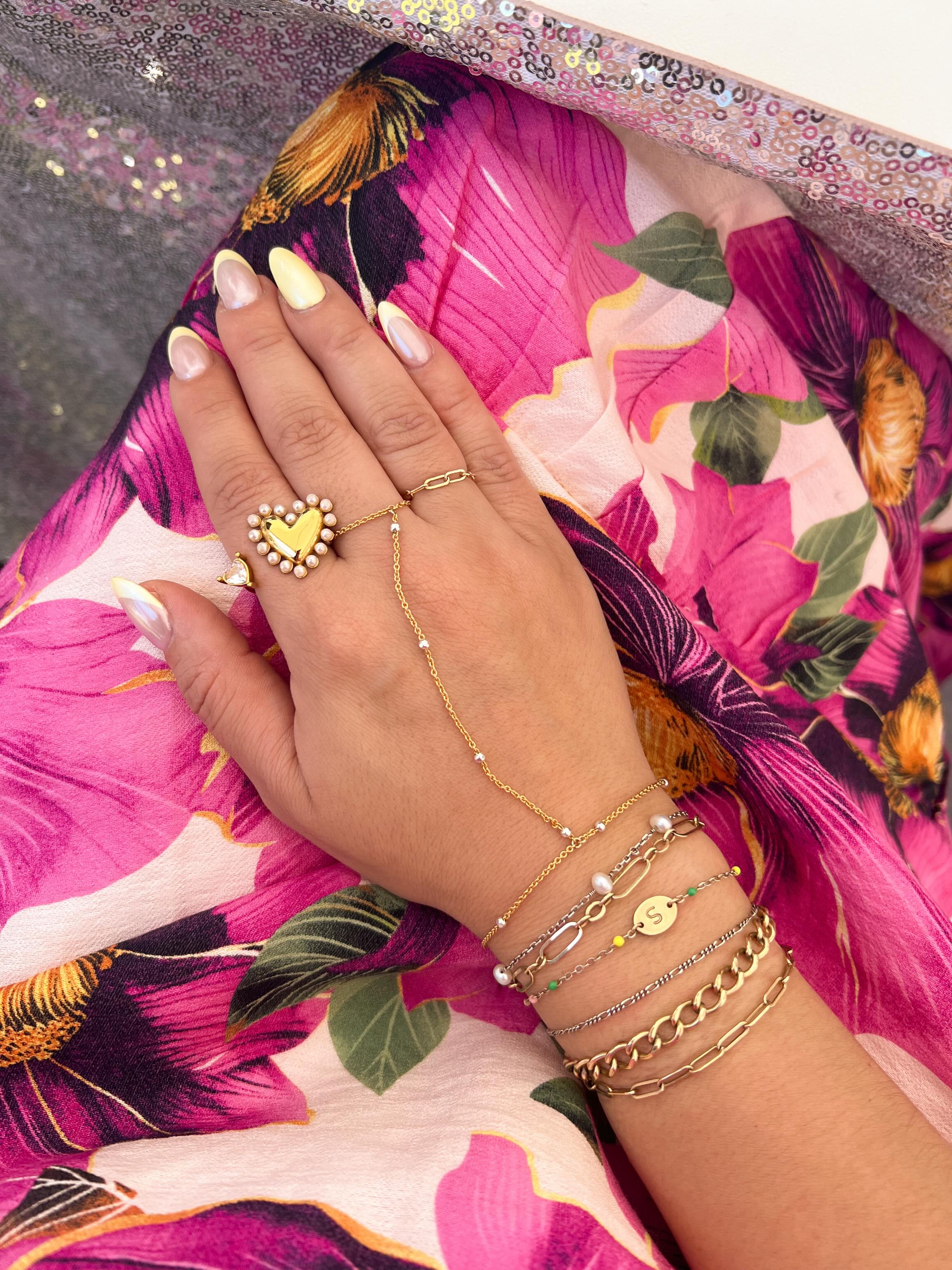Hand wearing gold hand chain and 14K gold filled and sterling silver permanent jewelry bracelets and rings with a colorful floral background