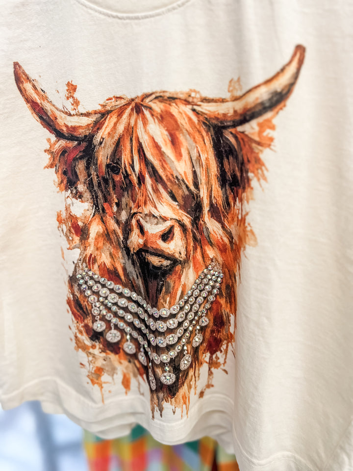 Cream t-shirt with a highland cow design wearing a rhinestone necklace on a cream background