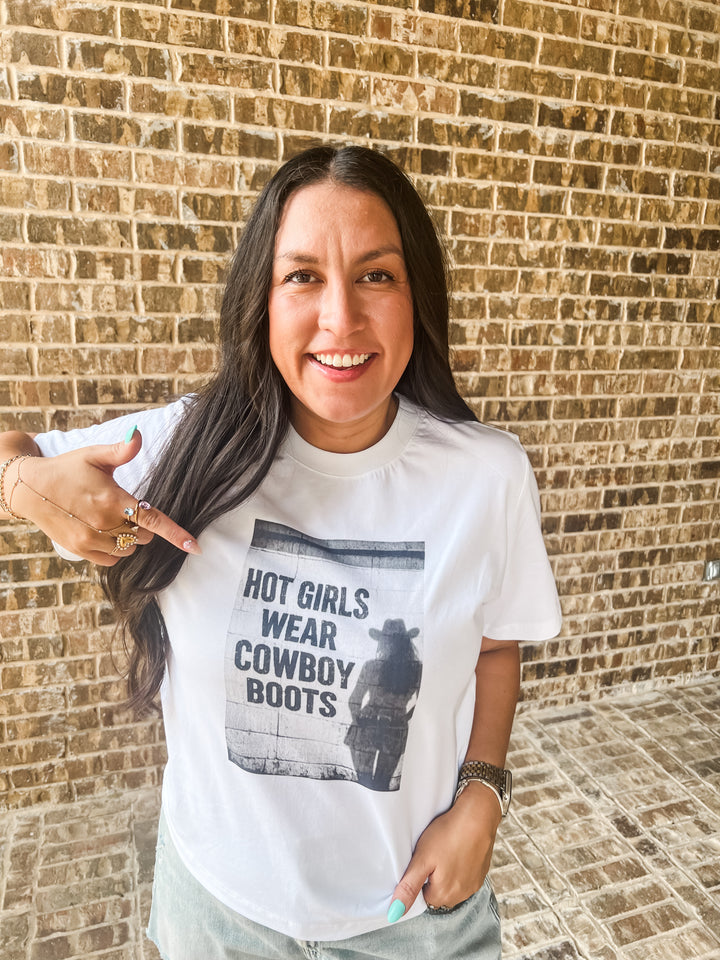 A woman with long dark hair stands in front of a tan brick wall, wearing a white cotton graphic T-shirt that reads &