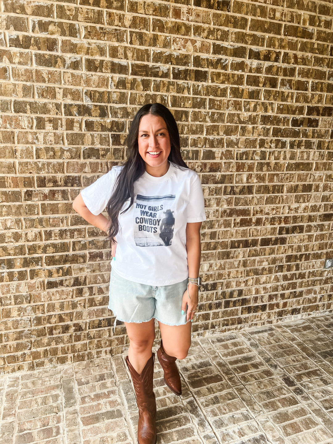 A woman posing in front of a brick wall wearing a white graphic t-shirt that reads &