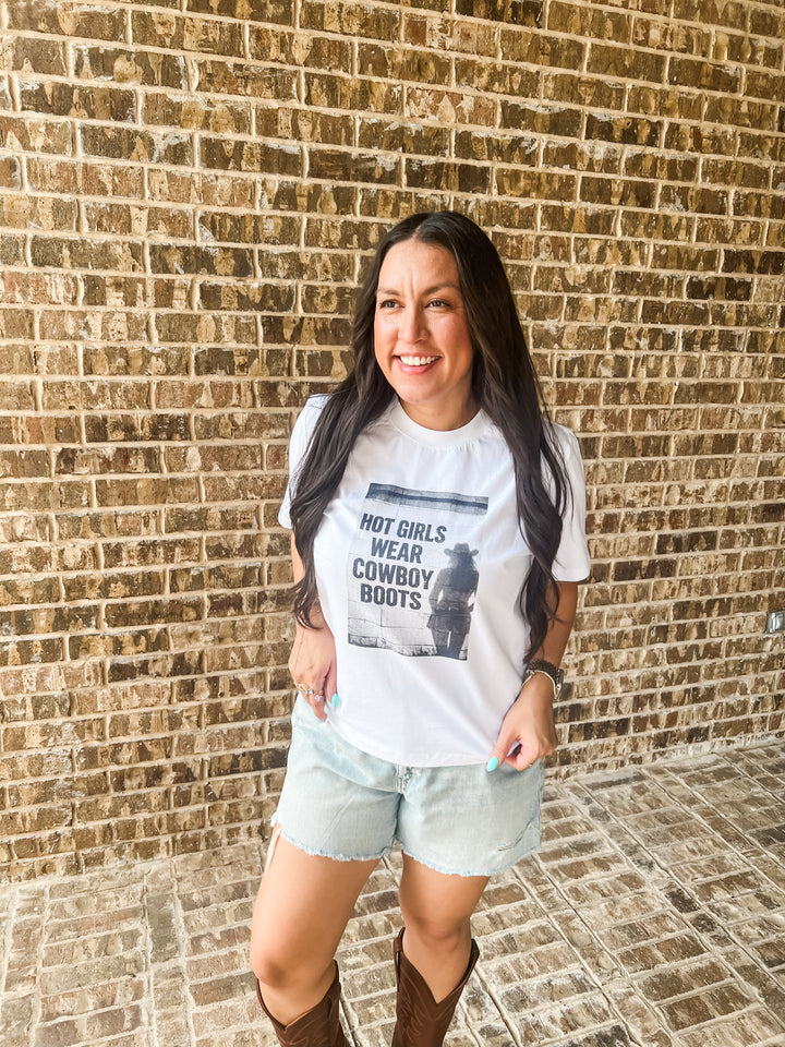 A woman posing in front of a brick wall wearing a white graphic t-shirt that reads &