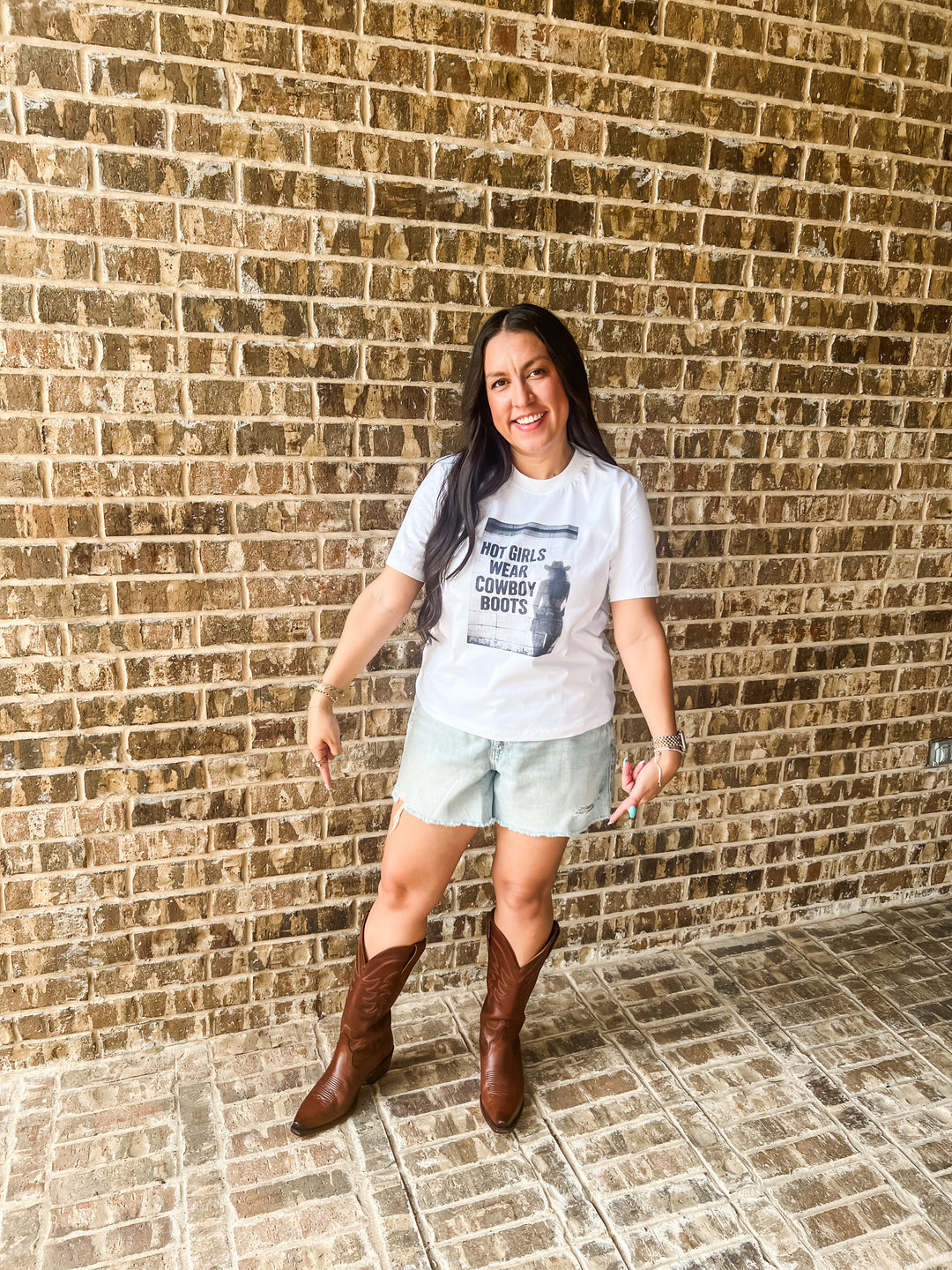 A woman posing in front of a brick wall wearing a white graphic t-shirt that reads &