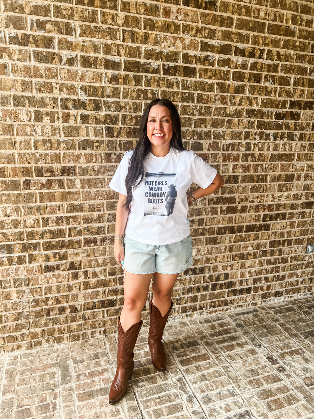A woman stands in front of a brick wall wearing a white graphic t-shirt that reads 'HOT GIRLS WEAR COWBOY BOOTS', light denim shorts, and brown cowboy boots.