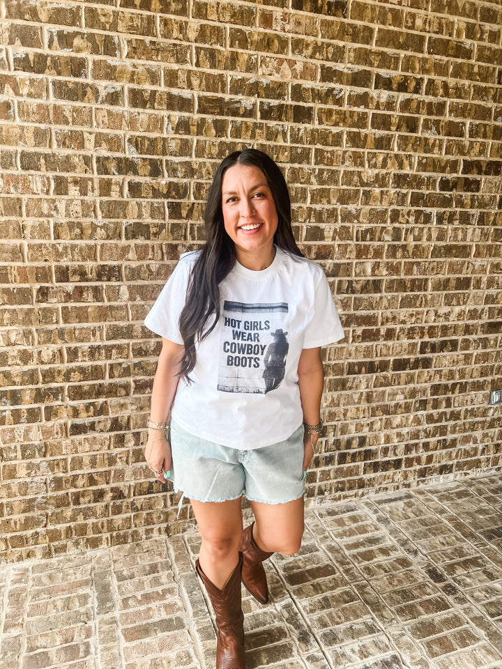 A woman stands in front of a brick wall wearing a white graphic t-shirt that reads &