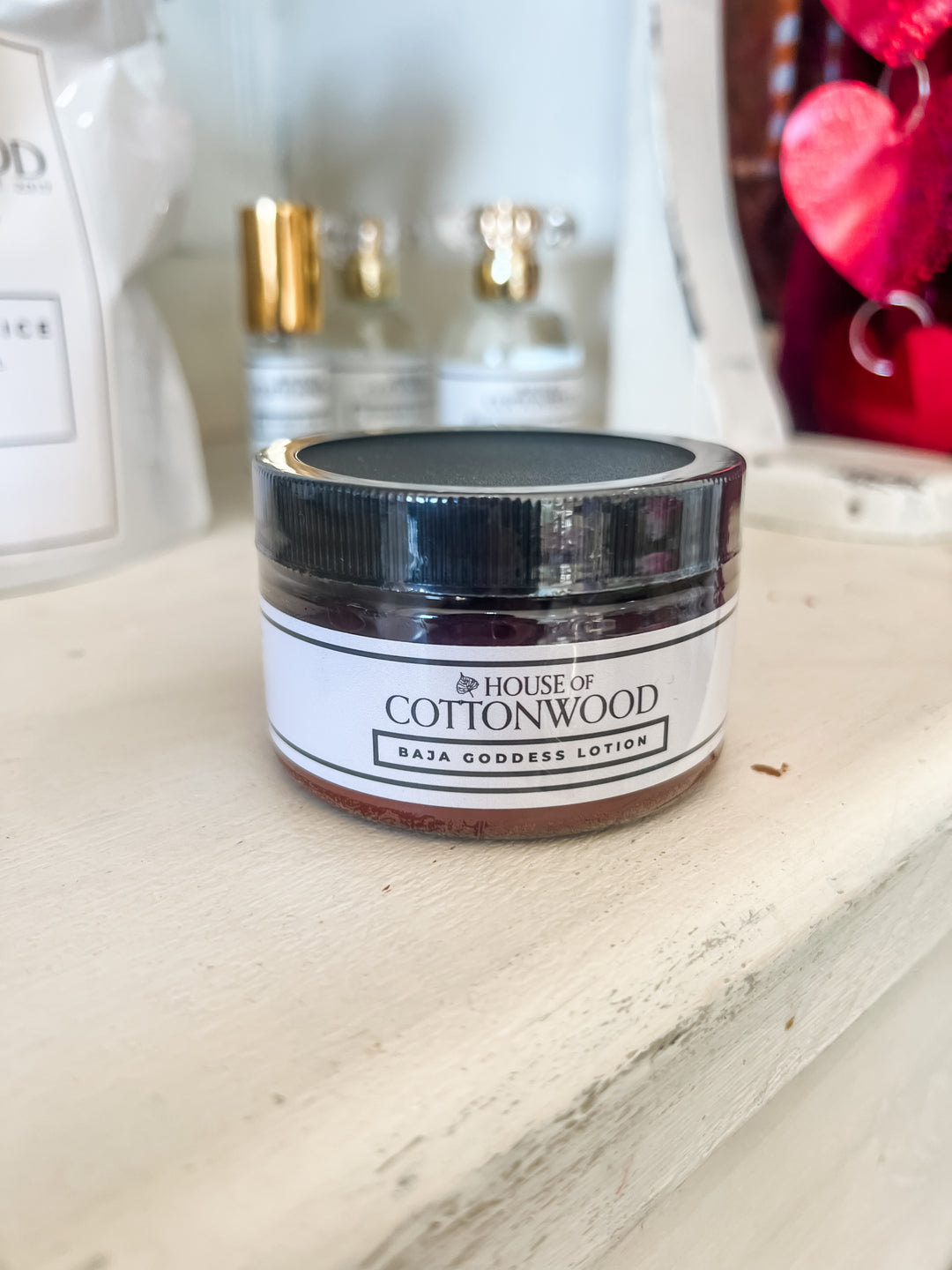 A jar of House of Cottonwood Baja Goddess Lotion sits on a distressed white wooden shelf, with other cosmetic products blurred in the background.