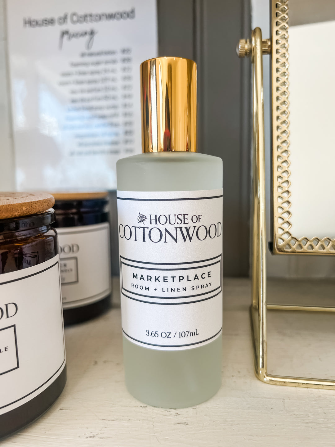 A frosted glass bottle of House of Cottonwood Marketplace room and linen spray with a metallic gold cap, displayed on a light wooden surface next to candles and a gold mirror frame.