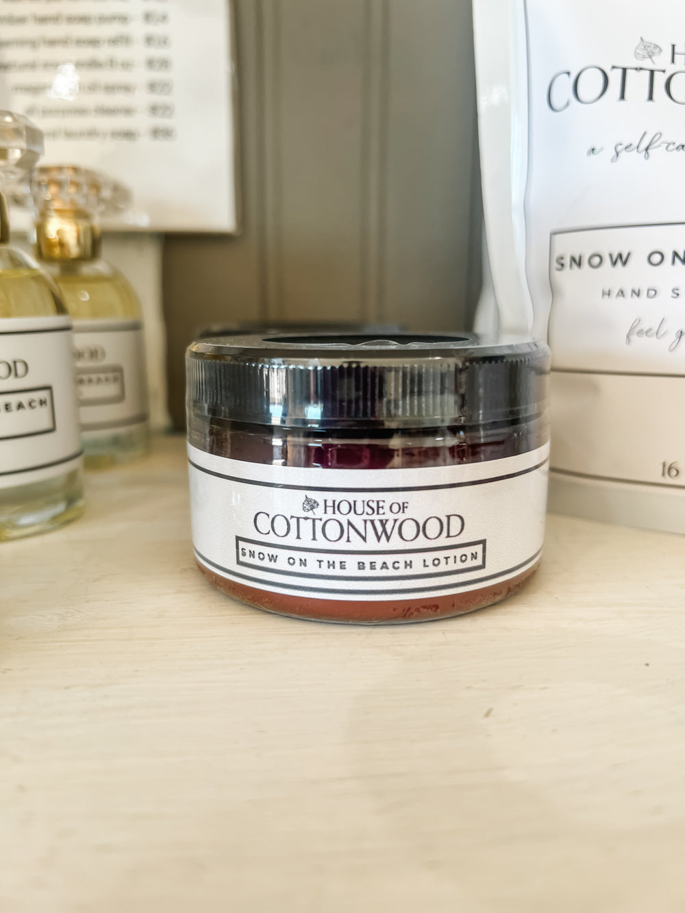 A small jar of House of Cottonwood Snow on the Beach Lotion with a black lid and white label, sitting on a light wooden surface with other products blurred in the background.