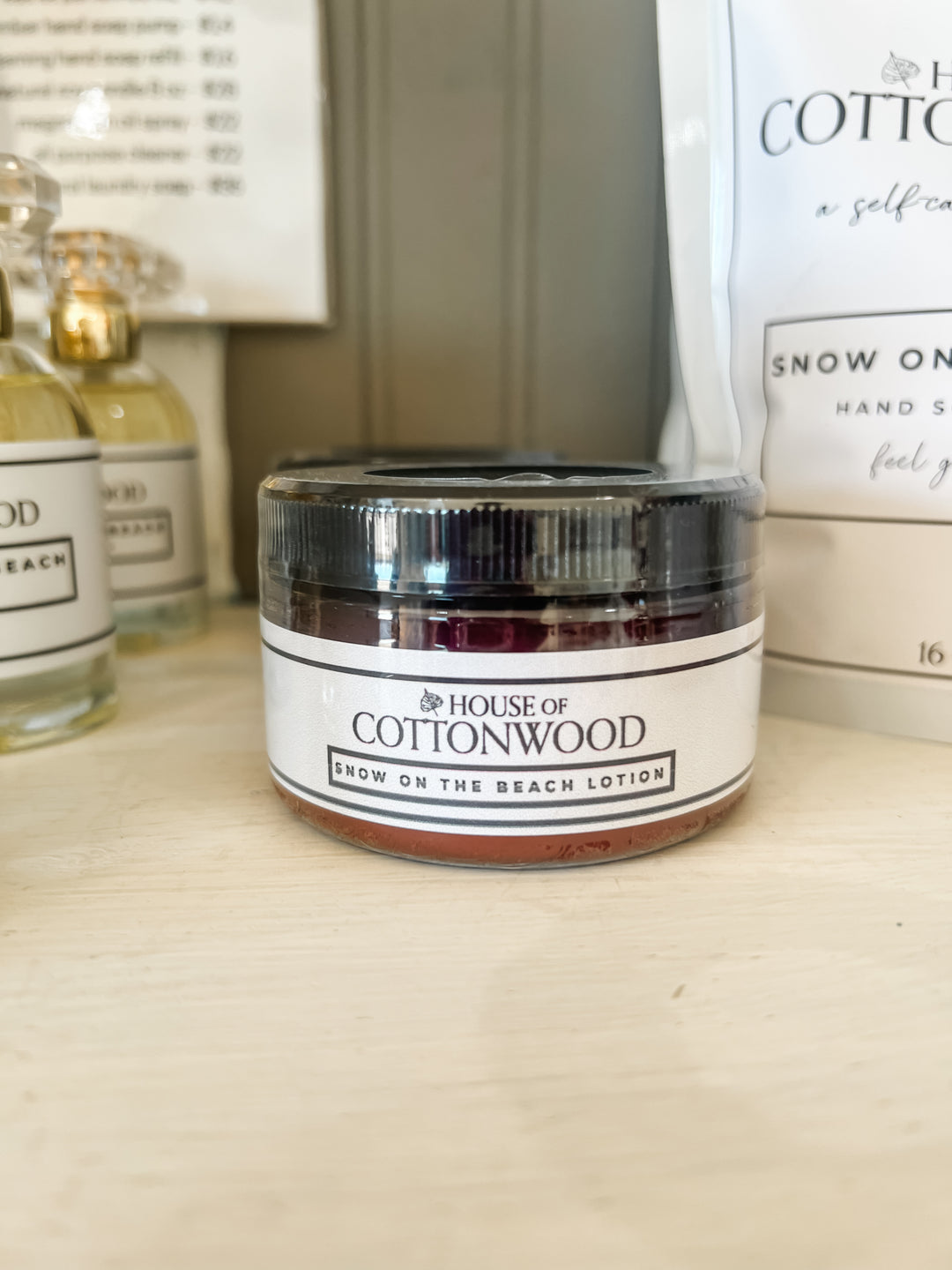 A small jar of House of Cottonwood Snow on the Beach Lotion with a black lid and white label, sitting on a light wooden surface with other products blurred in the background.