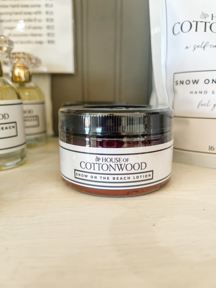 A small jar of House of Cottonwood Snow on the Beach Lotion with a black lid and white label, sitting on a light wooden surface with other products blurred in the background.