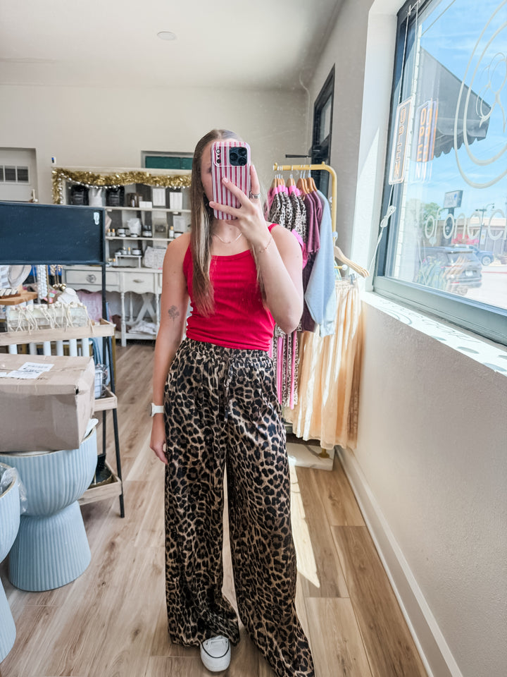 A woman takes a mirror selfie in a boutique, wearing a bright red tank top and high-waisted leopard-print wide-leg pants. The boutique interior features light wood floors, clothing racks, and a large window on the right.