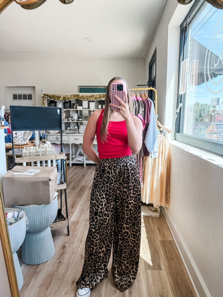 A woman takes a mirror selfie in a boutique, wearing a bright red tank top and wide-leg leopard print pants. The boutique has light wood floors, a clothing rack, and a window on the right.