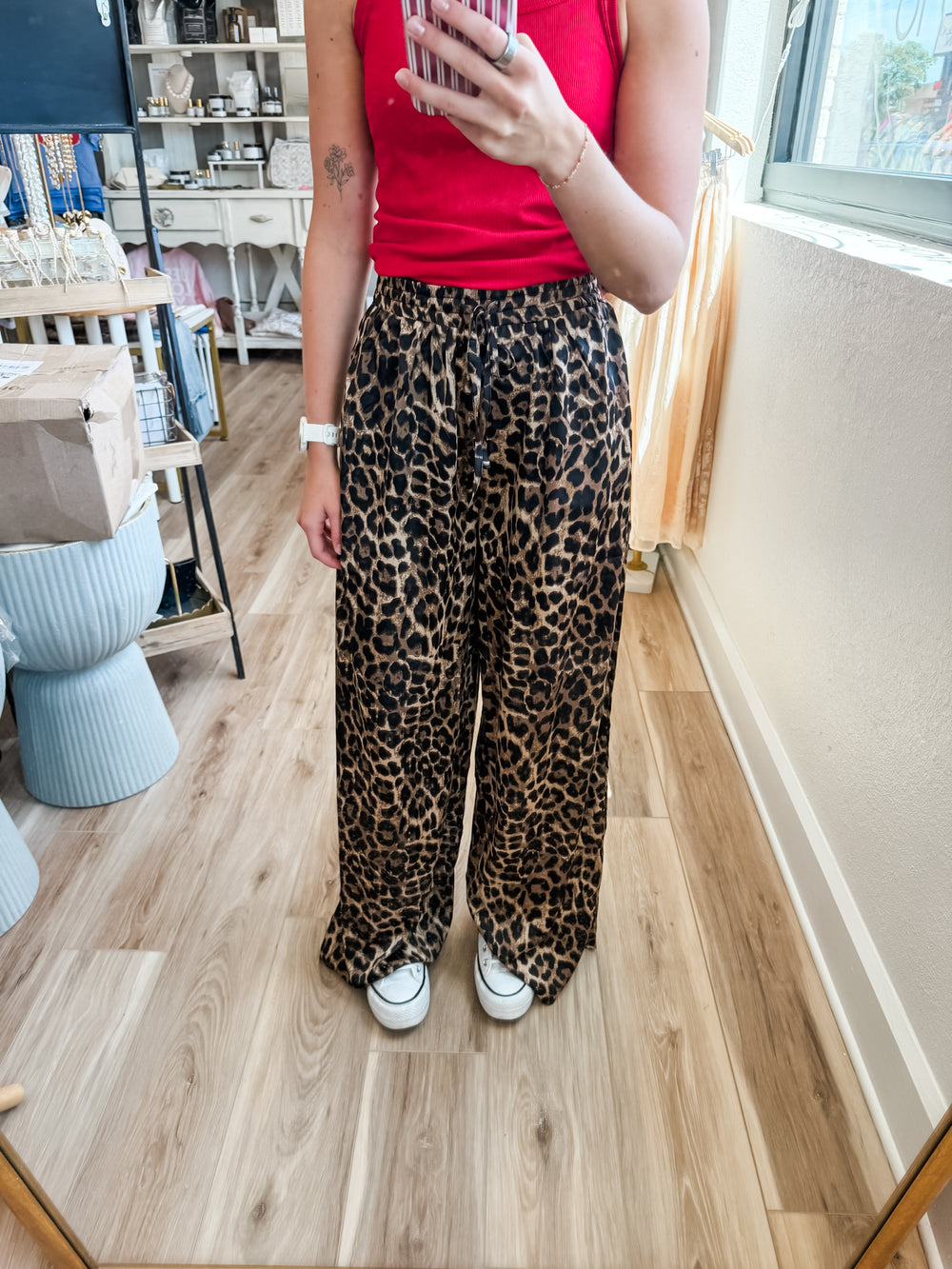 A person taking a mirror selfie in a boutique, wearing leopard print wide-leg pants, a red tank top, and white sneakers.