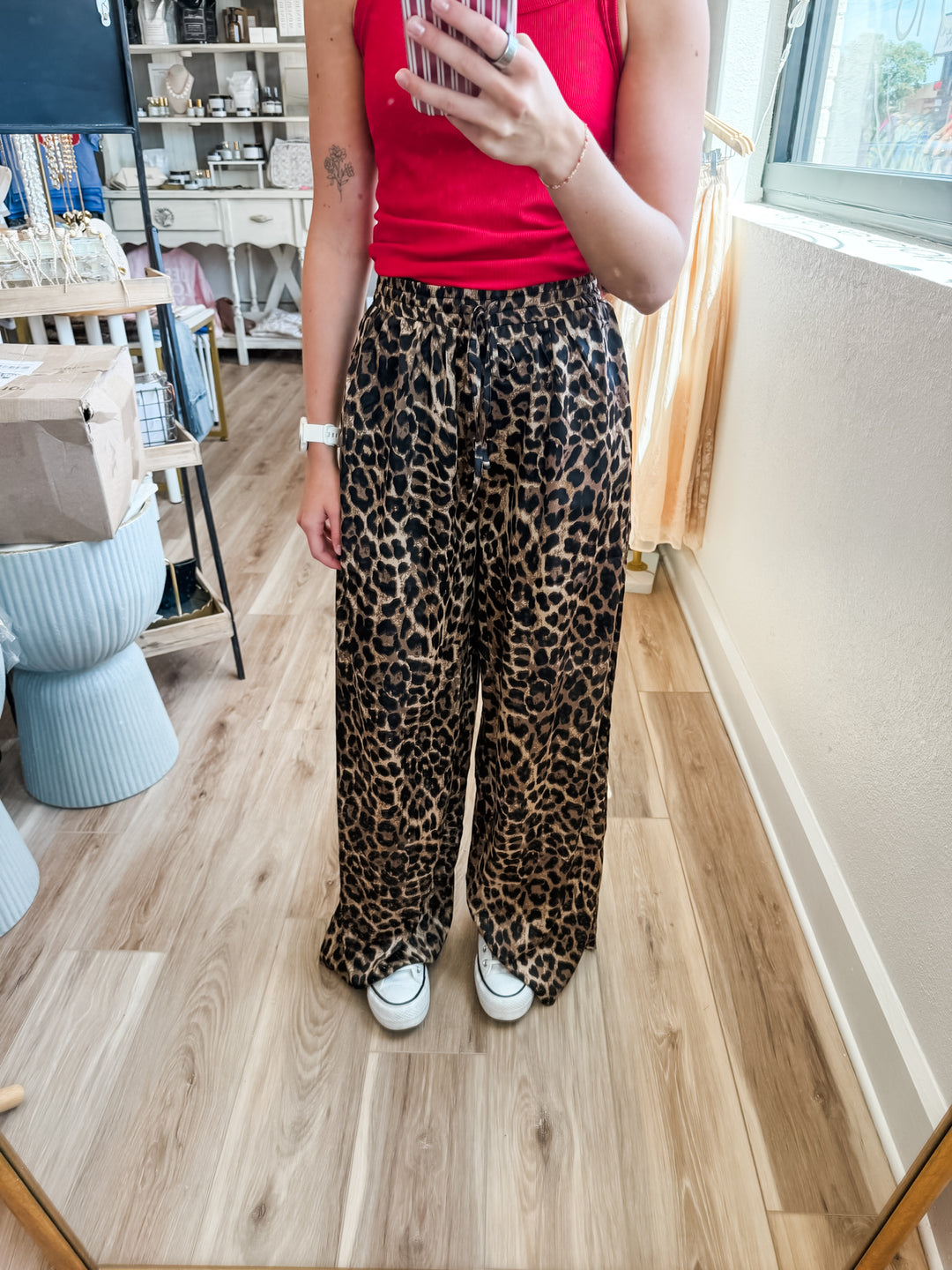 A person taking a mirror selfie in a boutique, wearing leopard print wide-leg pants, a red tank top, and white sneakers.