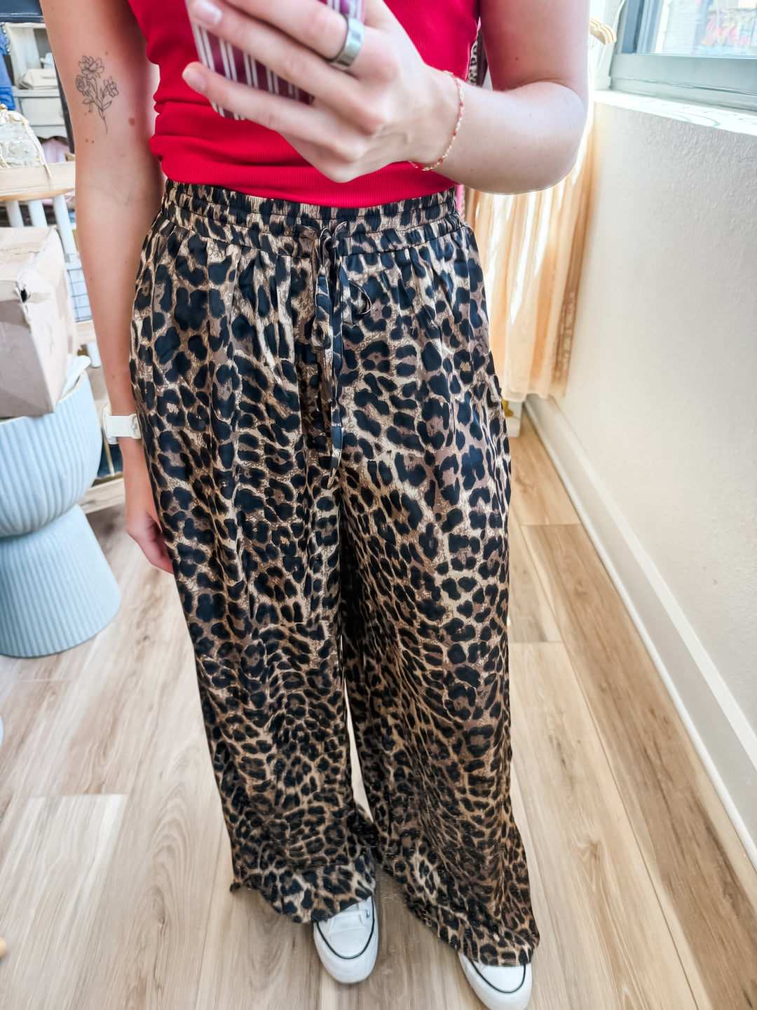 A person wearing loose, wide-leg leopard-print pants with a red top and white sneakers, standing indoors on a wood floor near a window.