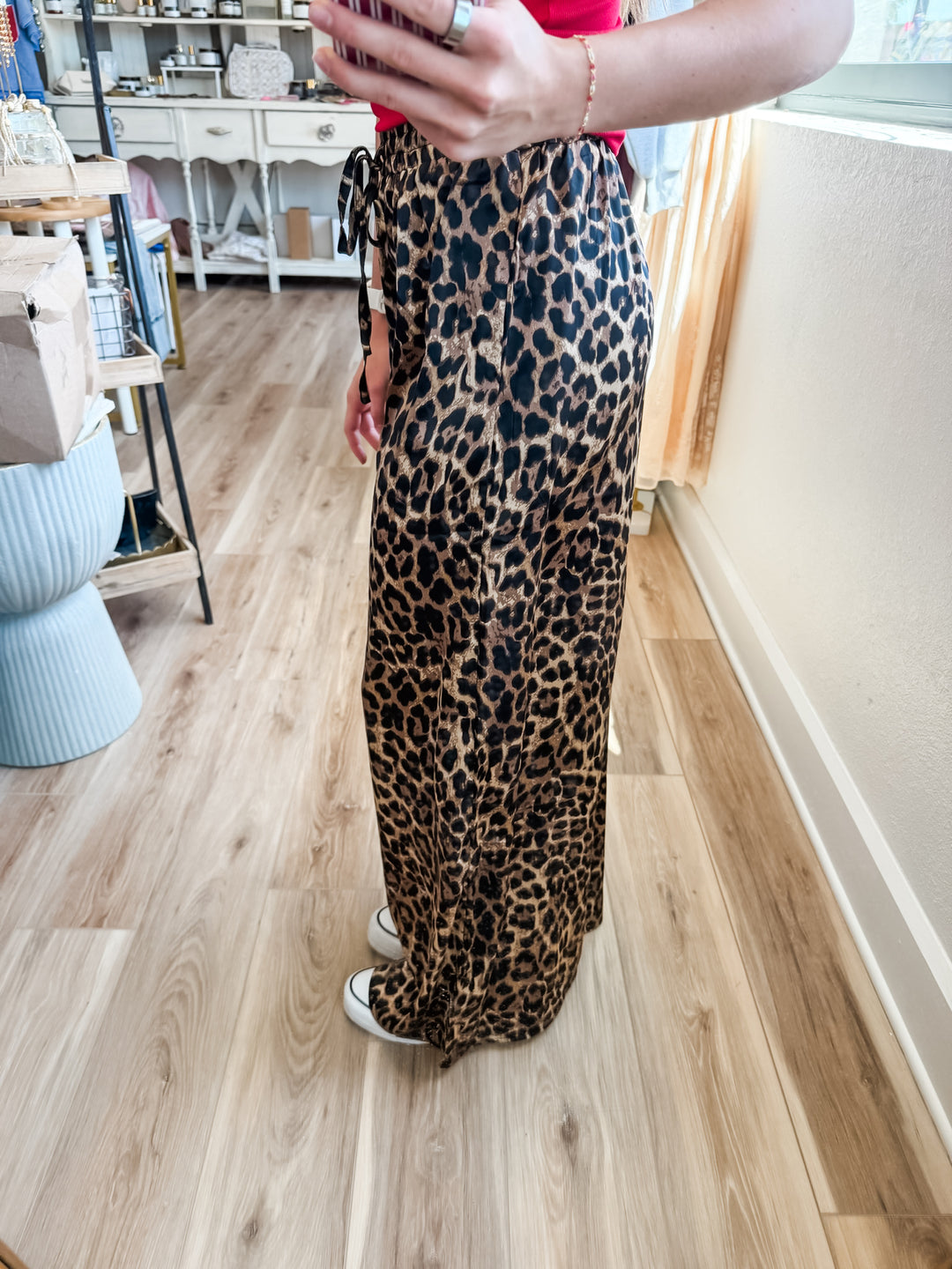 A person wearing leopard-print wide-leg pants and a red top, standing in a room with light wood floors and a window.