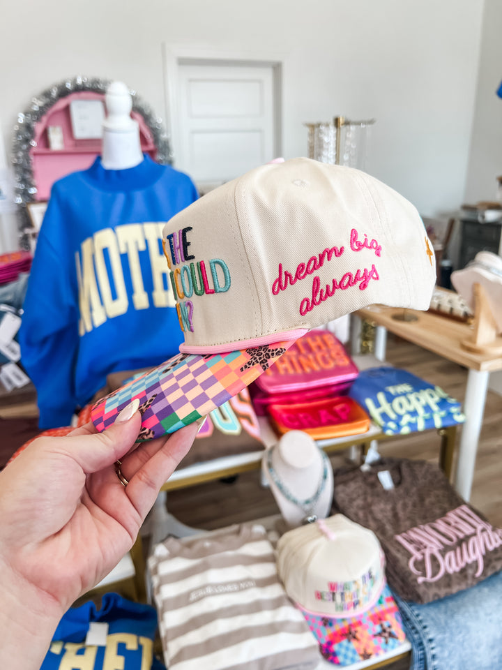 Beige cap with colorful text on the side "dream big always" held by a hand in a store setting 