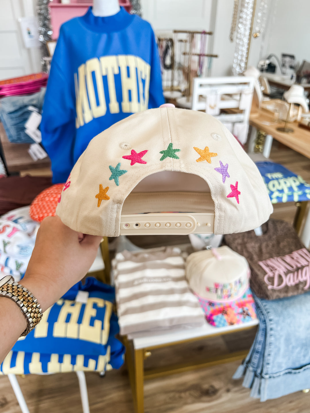 Beige cap with colorful star patterns on the back held by a person in a store setting.