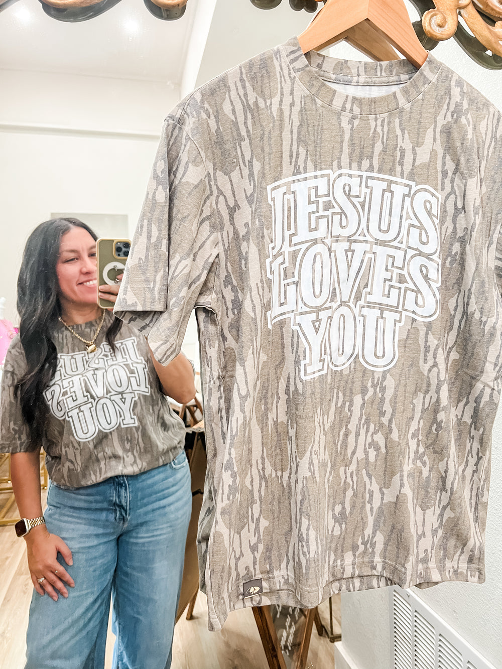 Person wearing a camouflage t-shirt with 'Jesus Loves You' text, standing next to another similar shirt on a hanger.