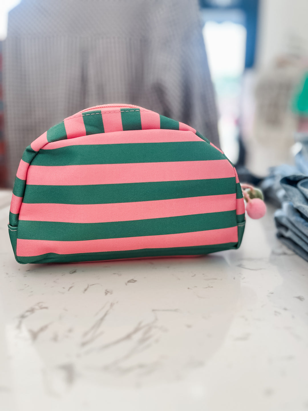 A small zippered cosmetic pouch with horizontal pink and dark green stripes, featuring a small pink pom-pom charm on the zipper, resting on a light marble surface.