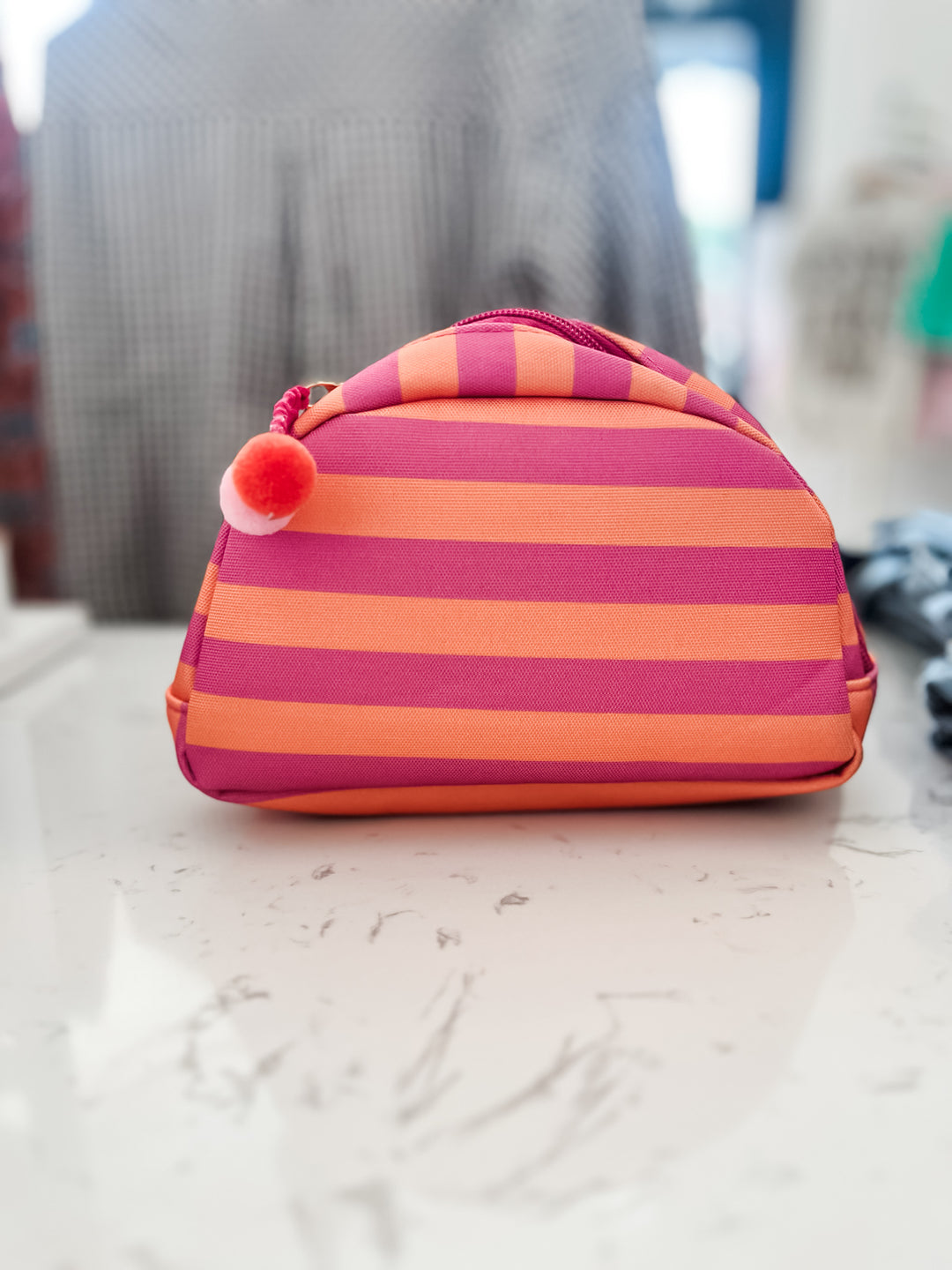 A small zippered cosmetic pouch with horizontal pink and orange stripes, featuring a red and white pom-pom charm on the zipper, sitting on a light marble surface.