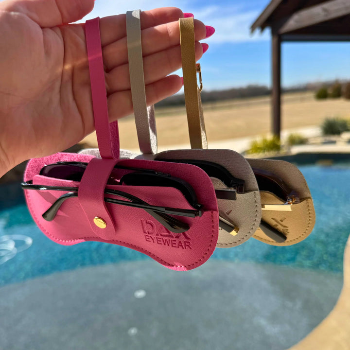 Three pairs of sunglasses in pink, beige, and gold cases held by a hand with a pool and outdoor setting in the background.